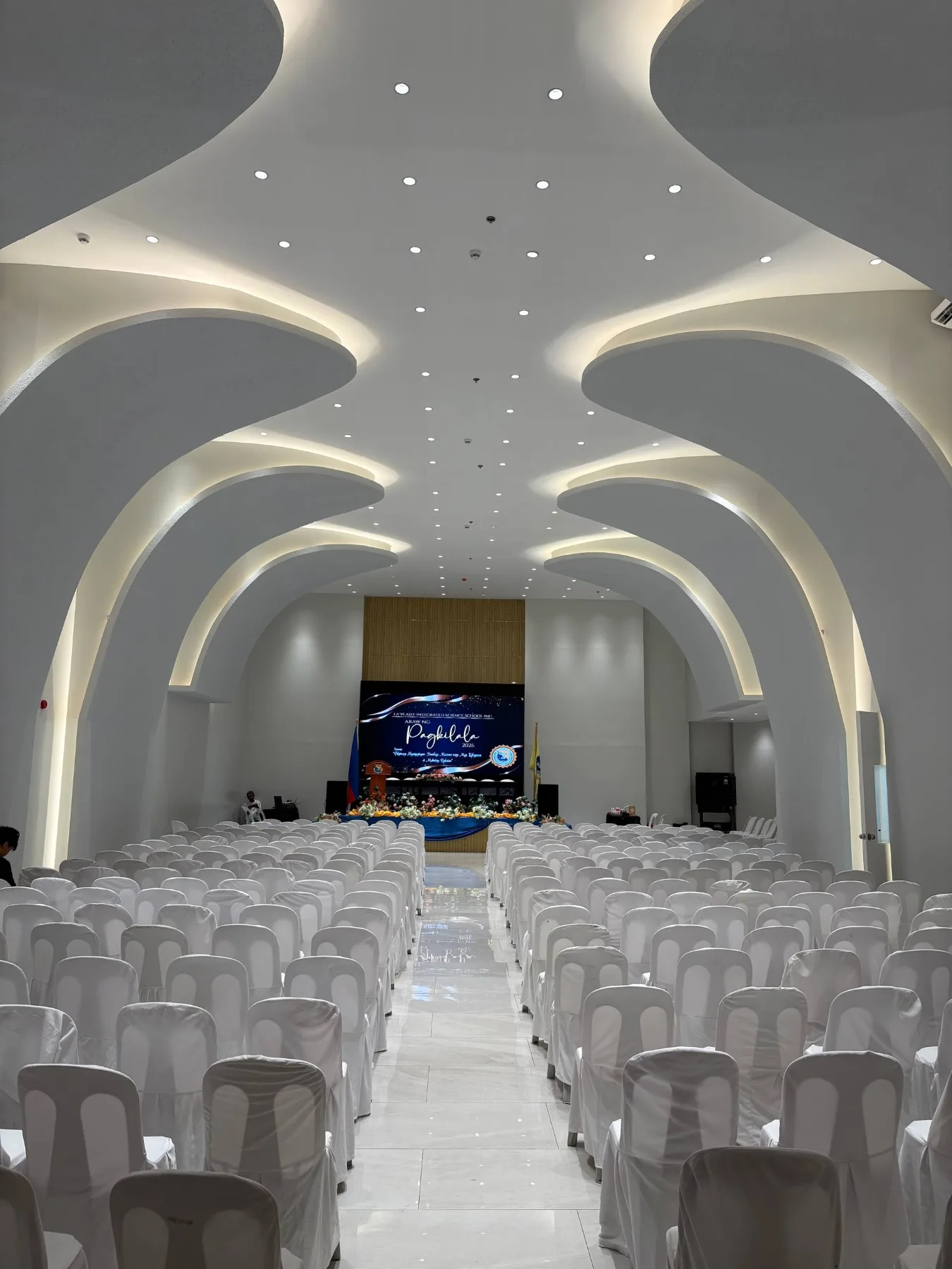 A grand central aisle leading up to a stage featuring a warm wood accent wall, floral arrangements, and a central graduation media display.